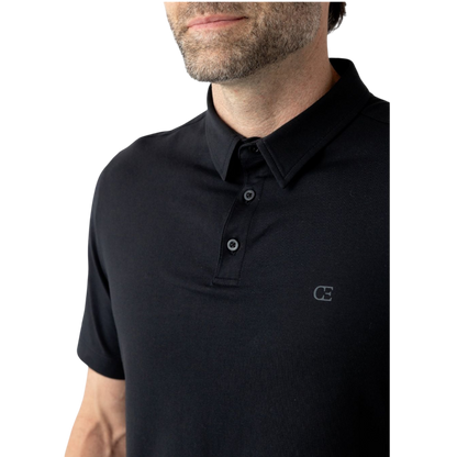 A person with short, light brown hair is wearing the Men's Everyday Polo by Cozy Earth in a light gray shade, paired with light beige pants. They are standing and slightly looking over their left shoulder with a neutral expression against a plain white background. |Color:Stone