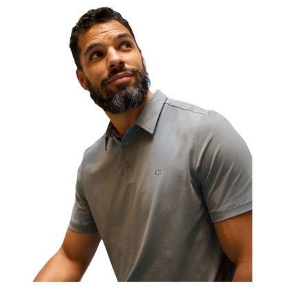 A person with short, light brown hair is wearing the Men's Everyday Polo by Cozy Earth in a light gray shade, paired with light beige pants. They are standing and slightly looking over their left shoulder with a neutral expression against a plain white background. |Color:Stone