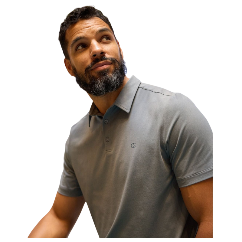 A person with short, light brown hair is wearing the Men's Everyday Polo by Cozy Earth in a light gray shade, paired with light beige pants. They are standing and slightly looking over their left shoulder with a neutral expression against a plain white background. |Color:Stone