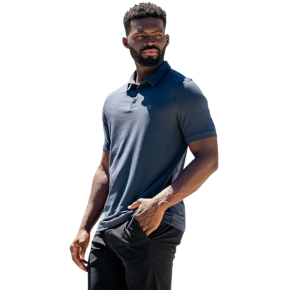 A person with short, light brown hair is wearing the Men's Everyday Polo by Cozy Earth in a light gray shade, paired with light beige pants. They are standing and slightly looking over their left shoulder with a neutral expression against a plain white background. |Color:Stone