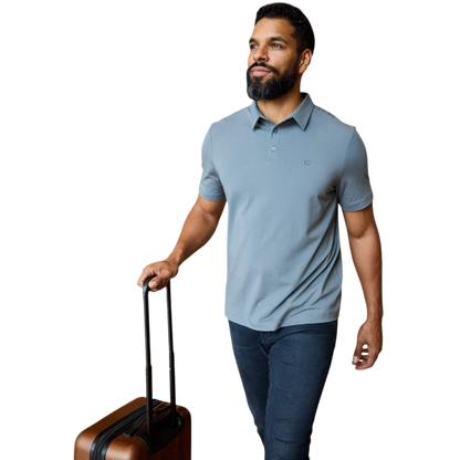 A person with short, light brown hair is wearing the Men's Everyday Polo by Cozy Earth in a light gray shade, paired with light beige pants. They are standing and slightly looking over their left shoulder with a neutral expression against a plain white background. |Color:Stone
