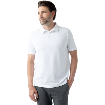 A man with short brown hair, a beard, and mustache is standing against a plain white background. He is wearing the Men's Everyday Polo from Cozy Earth in white and gray pants. One hand is in his pant pocket as he gazes to the side with a neutral expression. |Color:White