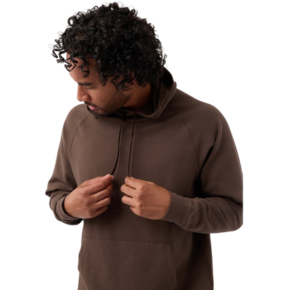 A person with curly hair is looking down and adjusting the drawstrings of their brown Men's StretchTech Hoodie from Cozy Earth. They are wearing a casual outfit and standing against a plain white background. |Color:Cacao