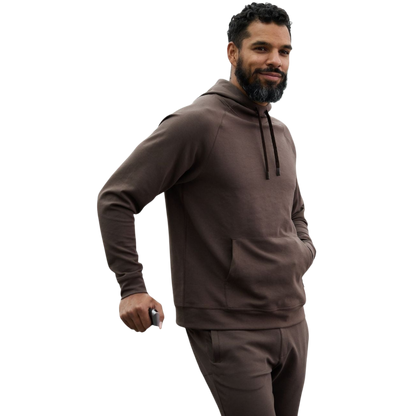 A person wearing the Men's StretchTech Hoodie from Cozy Earth, paired with matching gray pants, stands against a plain white background. The hoodie features drawstrings and a front pocket. The individual is looking forward with a neutral expression. |Color:Coal Heather