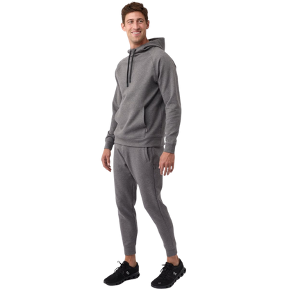 A person wearing the Men's StretchTech Hoodie from Cozy Earth, paired with matching gray pants, stands against a plain white background. The hoodie features drawstrings and a front pocket. The individual is looking forward with a neutral expression. |Color:Coal Heather