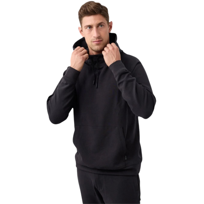 A man standing against a plain white background is wearing a black Men's StretchTech Hoodie and black pants from Cozy Earth. He is holding the hoodie strings with both hands. The man has short brown hair and a serious expression. |Color:Jet Black