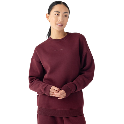 A person with dark hair tied back is wearing the Cozy Earth Women's CityScape Crewneck, paired with matching pants. They are standing against a plain white background. The crewneck features subtle lettering on the front. The person is smiling gently and has their hands clasped together. |Color:Burgundy