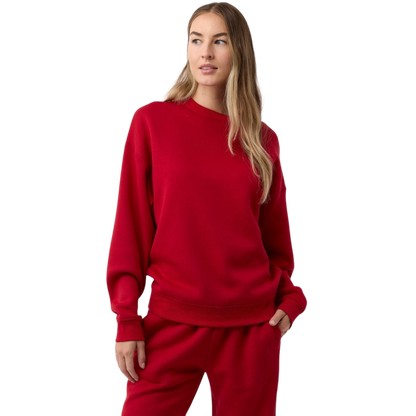 A person with long hair is wearing a Cozy Earth Women's CityScape Crewneck and matching sweatpants, standing against a plain white background. They have one hand in their pocket and are gazing slightly to the side. |Color:Crimson