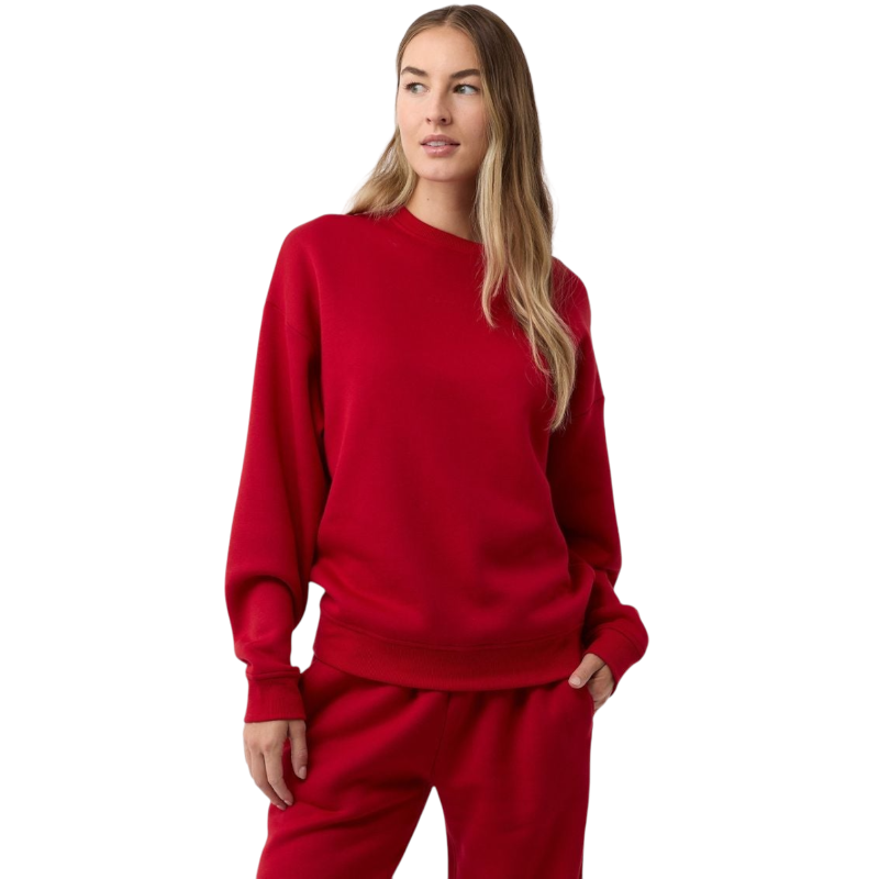 A person with long hair is wearing a Cozy Earth Women's CityScape Crewneck and matching sweatpants, standing against a plain white background. They have one hand in their pocket and are gazing slightly to the side. |Color:Crimson