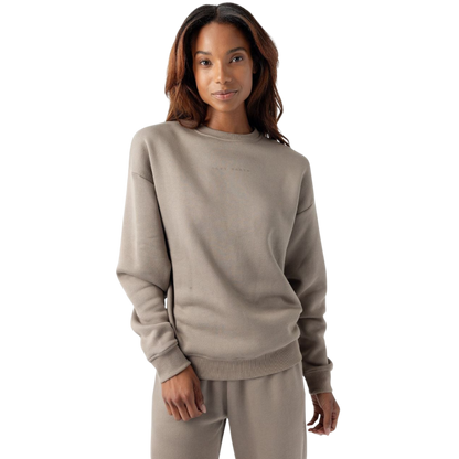 A person with long, wavy hair is wearing the Women's CityScape Crewneck and matching taupe-colored sweatpants from Cozy Earth. They are standing against a white background and smiling softly at the camera. The crewneck has subtle text on the chest. |Color:Ebony