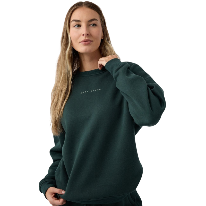 A person with long, light brown hair is wearing a dark green Women's CityScape Crewneck by Cozy Earth. They are looking slightly to the side, with one hand resting on the neckline of the sweatshirt. The background is plain white. |Color:Spruce