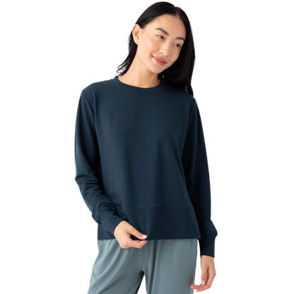 A person with long dark hair is wearing a dark blue Women's StudioLite Crewneck by Cozy Earth and light grey pants. They are standing against a plain white background, looking slightly to the side with a relaxed expression and holding the bottom of their top with one hand. |Color:Eclipse