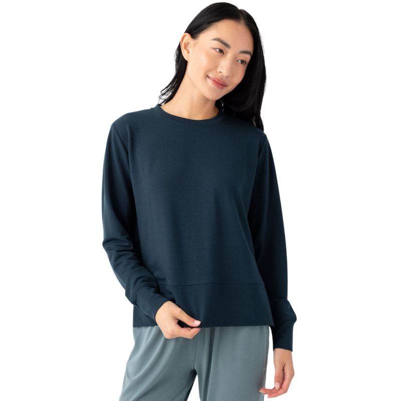 A person with long dark hair is wearing a dark blue Women's StudioLite Crewneck by Cozy Earth and light grey pants. They are standing against a plain white background, looking slightly to the side with a relaxed expression and holding the bottom of their top with one hand. |Color:Eclipse