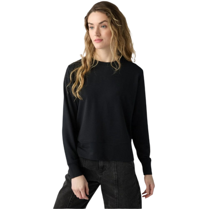 A woman with long wavy hair poses in a Cozy Earth Women's StudioLite Crewneck and dark jeans, with a neutral expression gazing slightly to the side against a simple white backdrop. |Color:Jet Black