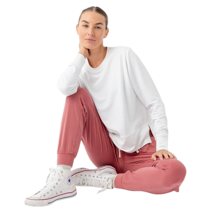 A person with long black hair is standing against a plain white background, wearing a Cozy Earth Women's StudioLite Crewneck in light green and dark green pants. They are looking down with a gentle smile, gently touching their neck with one hand. |Color:Haze