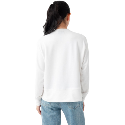A person with long black hair is standing against a plain white background, wearing a Cozy Earth Women's StudioLite Crewneck in light green and dark green pants. They are looking down with a gentle smile, gently touching their neck with one hand. |Color:Haze