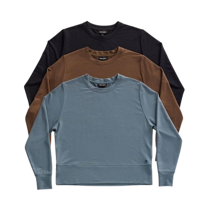 A person with long black hair is standing against a plain white background, wearing a Cozy Earth Women's StudioLite Crewneck in light green and dark green pants. They are looking down with a gentle smile, gently touching their neck with one hand. |Color:Haze
