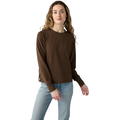 A person with long black hair is standing against a plain white background, wearing a Cozy Earth Women's StudioLite Crewneck in light green and dark green pants. They are looking down with a gentle smile, gently touching their neck with one hand. |Color:Haze