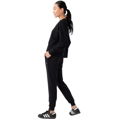 A person with long black hair is standing against a plain white background, wearing a Cozy Earth Women's StudioLite Crewneck in light green and dark green pants. They are looking down with a gentle smile, gently touching their neck with one hand. |Color:Haze