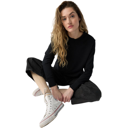 A person with long black hair is standing against a plain white background, wearing a Cozy Earth Women's StudioLite Crewneck in light green and dark green pants. They are looking down with a gentle smile, gently touching their neck with one hand. |Color:Haze