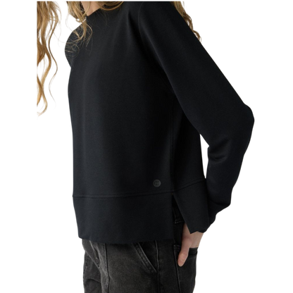 A person with long black hair is standing against a plain white background, wearing a Cozy Earth Women's StudioLite Crewneck in light green and dark green pants. They are looking down with a gentle smile, gently touching their neck with one hand. |Color:Haze