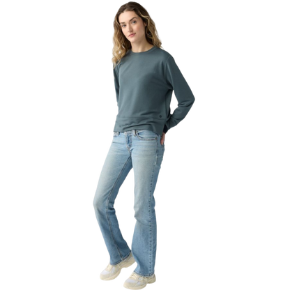 A person with long black hair is standing against a plain white background, wearing a Cozy Earth Women's StudioLite Crewneck in light green and dark green pants. They are looking down with a gentle smile, gently touching their neck with one hand. |Color:Haze