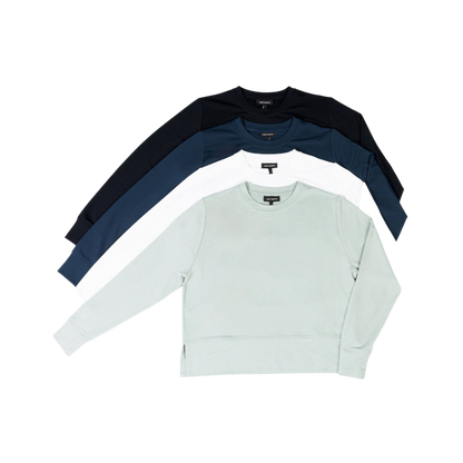 A person with long black hair is standing against a plain white background, wearing a Cozy Earth Women's StudioLite Crewneck in light green and dark green pants. They are looking down with a gentle smile, gently touching their neck with one hand. |Color:Haze