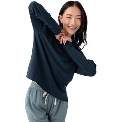 A person with long black hair is standing against a plain white background, wearing a Cozy Earth Women's StudioLite Crewneck in light green and dark green pants. They are looking down with a gentle smile, gently touching their neck with one hand. |Color:Haze