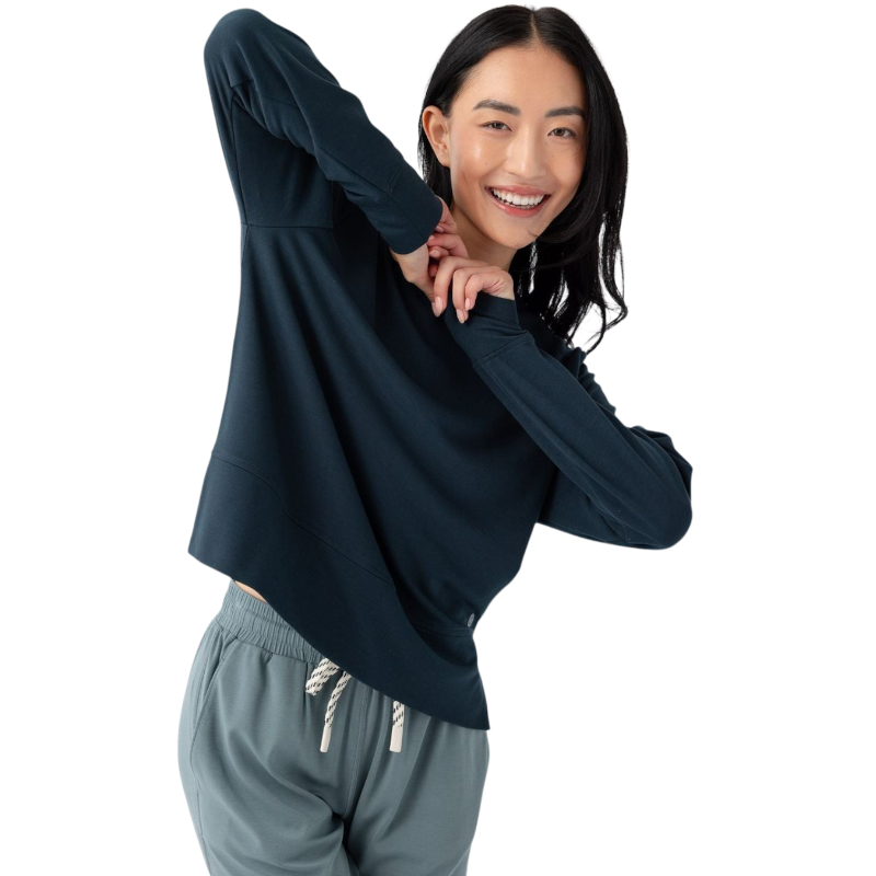 A person with long black hair is standing against a plain white background, wearing a Cozy Earth Women's StudioLite Crewneck in light green and dark green pants. They are looking down with a gentle smile, gently touching their neck with one hand. |Color:Haze