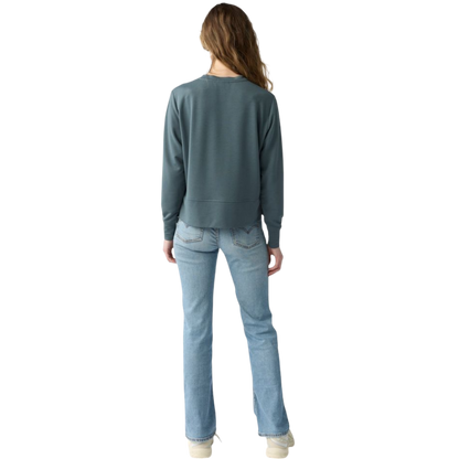 A person with long black hair is standing against a plain white background, wearing a Cozy Earth Women's StudioLite Crewneck in light green and dark green pants. They are looking down with a gentle smile, gently touching their neck with one hand. |Color:Haze