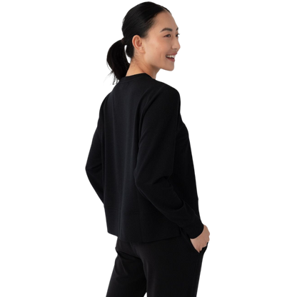 A person with long black hair is standing against a plain white background, wearing a Cozy Earth Women's StudioLite Crewneck in light green and dark green pants. They are looking down with a gentle smile, gently touching their neck with one hand. |Color:Haze