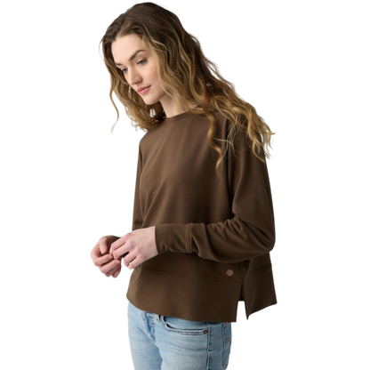 A person with long black hair is standing against a plain white background, wearing a Cozy Earth Women's StudioLite Crewneck in light green and dark green pants. They are looking down with a gentle smile, gently touching their neck with one hand. |Color:Haze