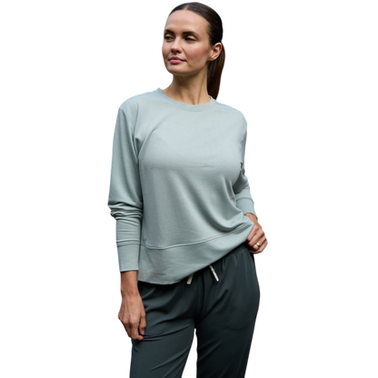 A person with long black hair is standing against a plain white background, wearing a Cozy Earth Women's StudioLite Crewneck in light green and dark green pants. They are looking down with a gentle smile, gently touching their neck with one hand. |Color:Haze