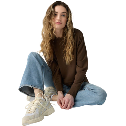 A person with long black hair is standing against a plain white background, wearing a Cozy Earth Women's StudioLite Crewneck in light green and dark green pants. They are looking down with a gentle smile, gently touching their neck with one hand. |Color:Haze