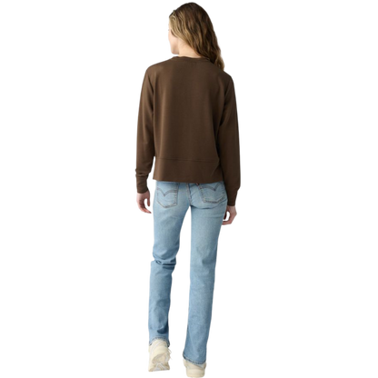 A person with long black hair is standing against a plain white background, wearing a Cozy Earth Women's StudioLite Crewneck in light green and dark green pants. They are looking down with a gentle smile, gently touching their neck with one hand. |Color:Haze