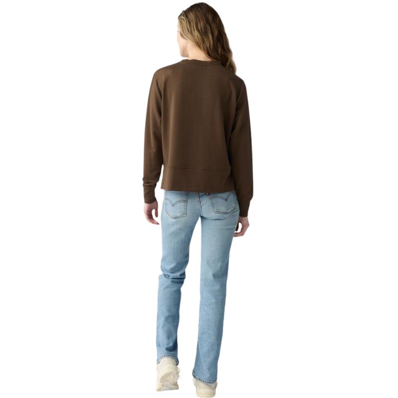 A person with long black hair is standing against a plain white background, wearing a Cozy Earth Women's StudioLite Crewneck in light green and dark green pants. They are looking down with a gentle smile, gently touching their neck with one hand. |Color:Haze