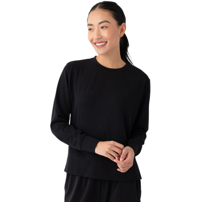 A person with long black hair is standing against a plain white background, wearing a Cozy Earth Women's StudioLite Crewneck in light green and dark green pants. They are looking down with a gentle smile, gently touching their neck with one hand. |Color:Haze