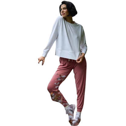 A person with long black hair is standing against a plain white background, wearing a Cozy Earth Women's StudioLite Crewneck in light green and dark green pants. They are looking down with a gentle smile, gently touching their neck with one hand. |Color:Haze
