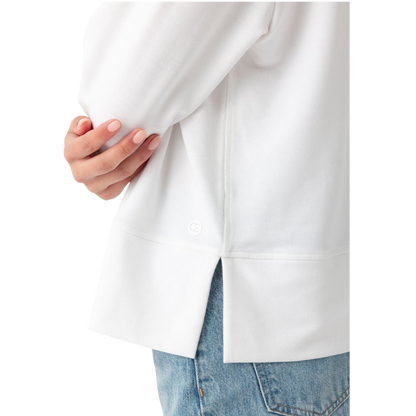 A person with long black hair is standing against a plain white background, wearing a Cozy Earth Women's StudioLite Crewneck in light green and dark green pants. They are looking down with a gentle smile, gently touching their neck with one hand. |Color:Haze