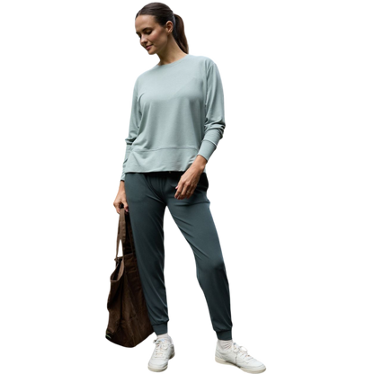 A person with long black hair is standing against a plain white background, wearing a Cozy Earth Women's StudioLite Crewneck in light green and dark green pants. They are looking down with a gentle smile, gently touching their neck with one hand. |Color:Haze
