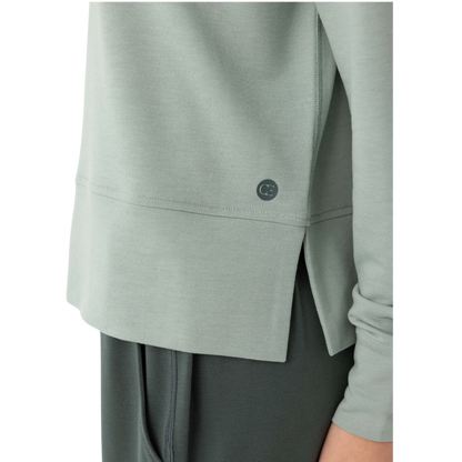 A person with long black hair is standing against a plain white background, wearing a Cozy Earth Women's StudioLite Crewneck in light green and dark green pants. They are looking down with a gentle smile, gently touching their neck with one hand. |Color:Haze