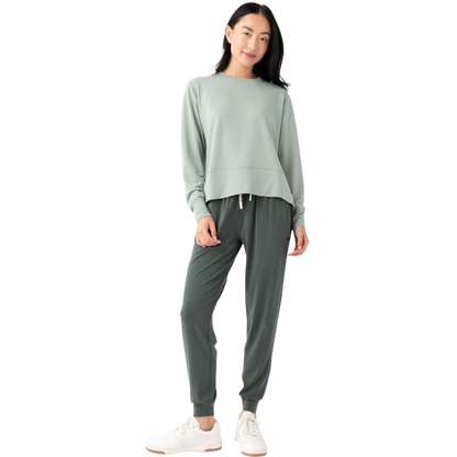 A person with long black hair is standing against a plain white background, wearing a Cozy Earth Women's StudioLite Crewneck in light green and dark green pants. They are looking down with a gentle smile, gently touching their neck with one hand. |Color:Haze