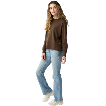 A person with long black hair is standing against a plain white background, wearing a Cozy Earth Women's StudioLite Crewneck in light green and dark green pants. They are looking down with a gentle smile, gently touching their neck with one hand. |Color:Haze