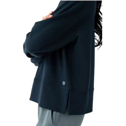 A person with long black hair is standing against a plain white background, wearing a Cozy Earth Women's StudioLite Crewneck in light green and dark green pants. They are looking down with a gentle smile, gently touching their neck with one hand. |Color:Haze