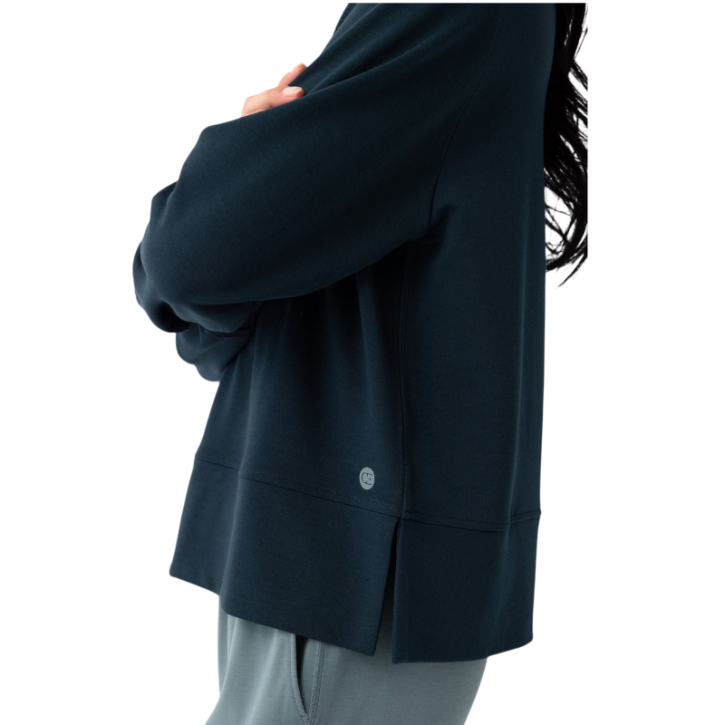 A person with long black hair is standing against a plain white background, wearing a Cozy Earth Women's StudioLite Crewneck in light green and dark green pants. They are looking down with a gentle smile, gently touching their neck with one hand. |Color:Haze