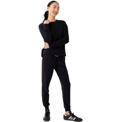 A person with long black hair is standing against a plain white background, wearing a Cozy Earth Women's StudioLite Crewneck in light green and dark green pants. They are looking down with a gentle smile, gently touching their neck with one hand. |Color:Haze