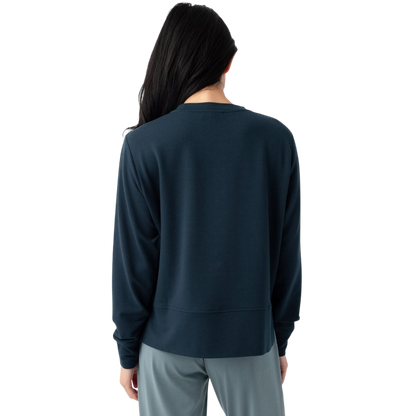 A person with long black hair is standing against a plain white background, wearing a Cozy Earth Women's StudioLite Crewneck in light green and dark green pants. They are looking down with a gentle smile, gently touching their neck with one hand. |Color:Haze