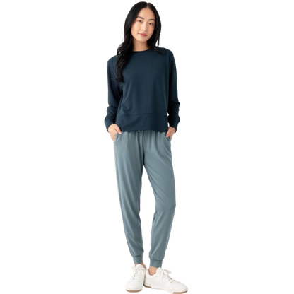 A person with long black hair is standing against a plain white background, wearing a Cozy Earth Women's StudioLite Crewneck in light green and dark green pants. They are looking down with a gentle smile, gently touching their neck with one hand. |Color:Haze