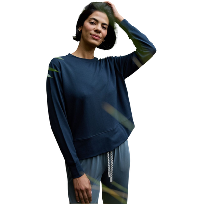 A person with long black hair is standing against a plain white background, wearing a Cozy Earth Women's StudioLite Crewneck in light green and dark green pants. They are looking down with a gentle smile, gently touching their neck with one hand. |Color:Haze