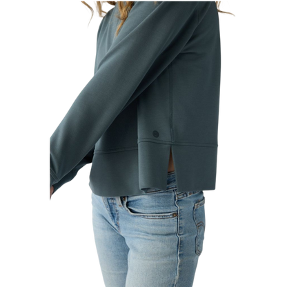 A person with long black hair is standing against a plain white background, wearing a Cozy Earth Women's StudioLite Crewneck in light green and dark green pants. They are looking down with a gentle smile, gently touching their neck with one hand. |Color:Haze
